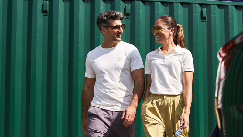 Hombre con playera blanca y mujer con polo blanco caminan frente a contenedores verdes.