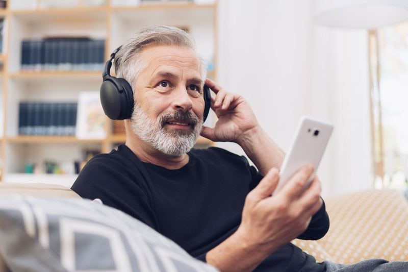 Mann mittleren Alters sitzt auf einer Couch und lauscht über seine schwarzen Kopfhörer interessiert einem Podcast. 