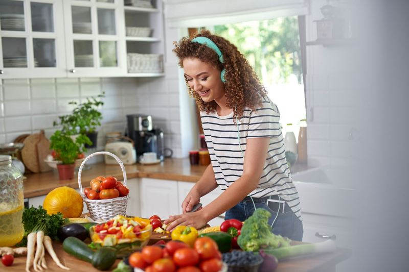 Eine junge Frau mit Kopfhörern auf den Ohren schneidet Gemüse in einer hellen Küche. 