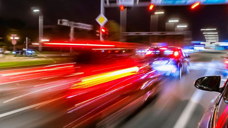 Ein Auto bremst bei Nacht an einer roten Ampel. Bewegungsunschärfe lässt die Rücklichter Streifen ziehen