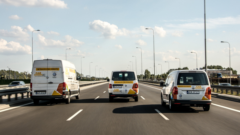 Dostawcze Volkswageny z okleiną w ofercie carsharingu jadą po trójpasmowej ulicy.
