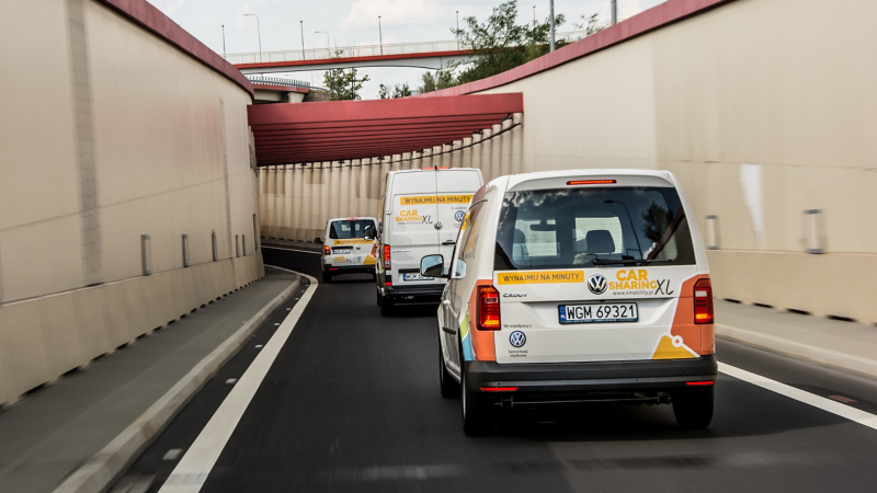 Dostawcze Volkswageny z okleiną w ofercie carsharingu jadą po ulicy.