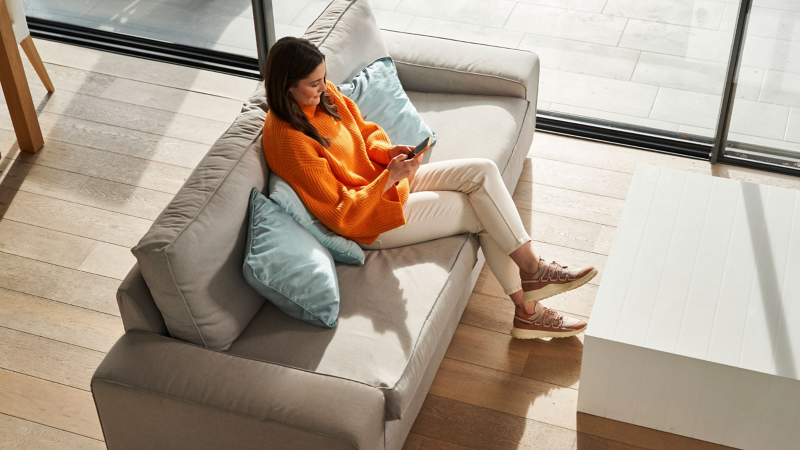 Mujer con celular en las manos sentada en sofa, dentro de habitación iluminada con luz del atardecer.