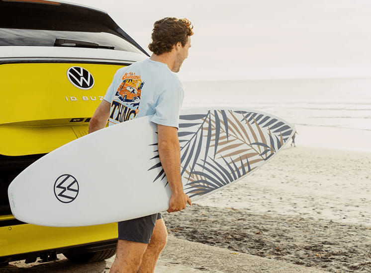 A person walks along the beach, carrying a surfboard adorned with a palm leaf design. Behind them, the vibrant Volkswagen ID Buzz stands out