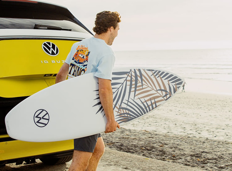 Man with surfboard walks to ocean, Volkswagen ID. Buzz nearby.