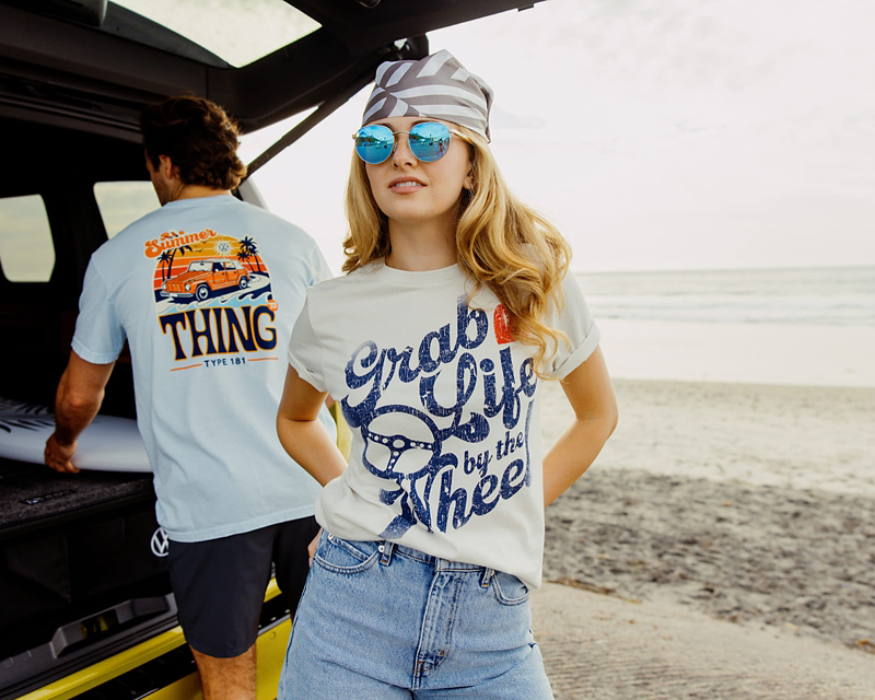 Mujer joven con camiseta "Grab Life by the Wheel" en la playa, hombre con tabla de surf.