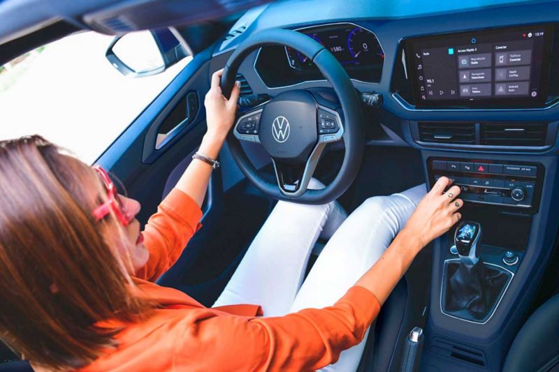 Mujer con blusa naranja y pantalón blanco maneja un SUV T Cross de Volkswagen, versión Trendline.