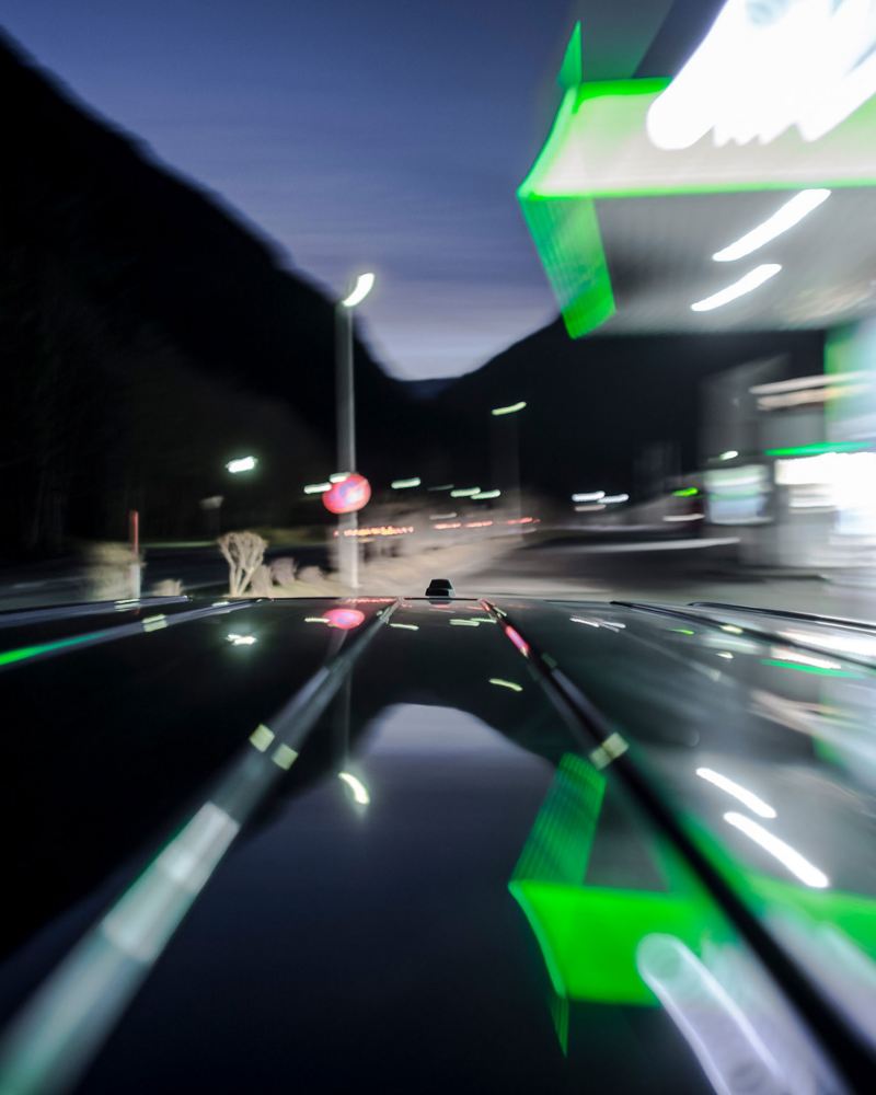 Langzeitbelichtung einer beleuchteten Tankstelle bei Nacht, vom Dach eines fahrenden Autos