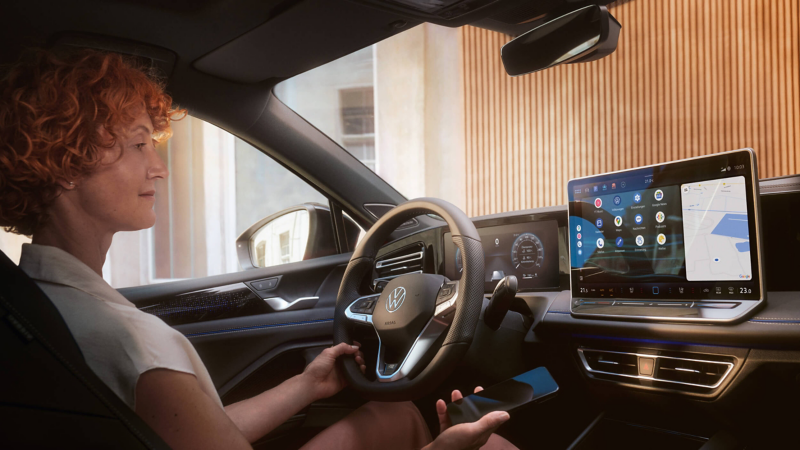A woman sitting in the driver's seat of a parked car, looking at the multimedia display, and with a smartphone in her hand
