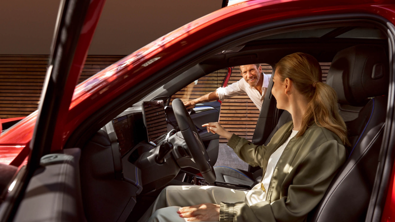 A woman sitting in the driver's seat of a VW with a man leaning in through the open passenger-side door