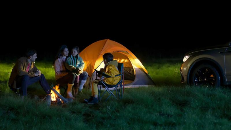 Familia acampa en el bosque, iluminados por los faros de SUV 2024 Tiguan Volkswagen.
