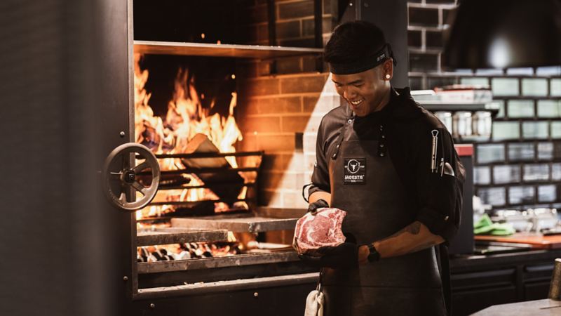 Ein Koch betrachtet ein Stück Fleisch vor einem offenen Feuer.