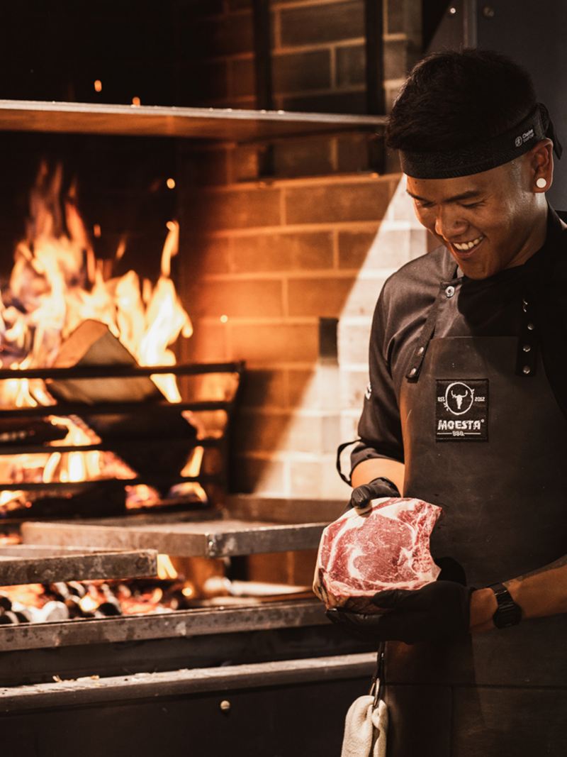 Ein Koch betrachtet ein Stück Fleisch vor einem offenen Feuer.