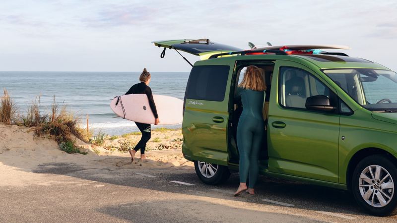 Två kvinnor packar ur surfingbrädor ur en grön VW Caddy California campingbil på stranden