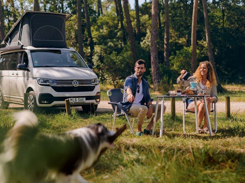 VW California med popup-taket uppe och två personer med hund som fikar vid det hopfällbara campingbordet