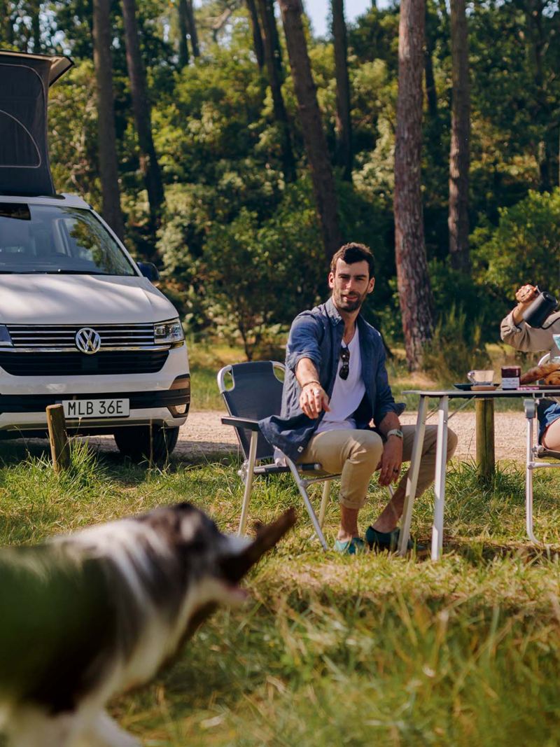 VW California med popup-taket uppe och två personer med hund som fikar vid det hopfällbara campingbordet