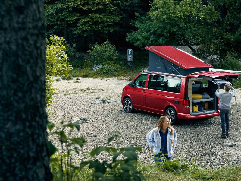 Ett par packar ur en röd VW California campingbuss på en stenig strand vid en sjö