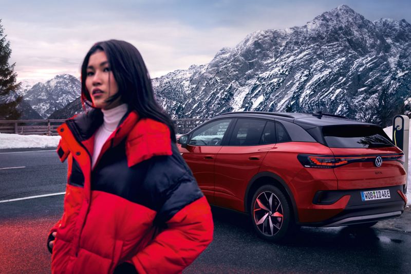: Eine Frau steht vor einem Volkswagen ID. 4 an einer Straße durch verschneite Berge.