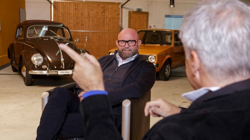 Claude Gregorini hört Dino Graf zu und sitzt auf einem Sessel vor einem alten VW Käfer
