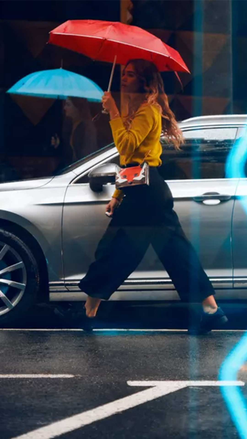 Une femme aux cheveux longs marche dans la rue sous un parapluie rouge, vêtue d'un haut jaune et d'un pantalon foncé, avec une voiture VW argentée visible en arrière-plan. Un deuxième parapluie bleu est flou en arrière-plan.
