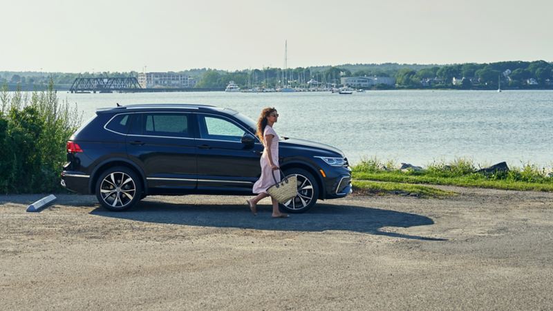 A woman walks by a 2024 blue VW Tiguan parked by the water