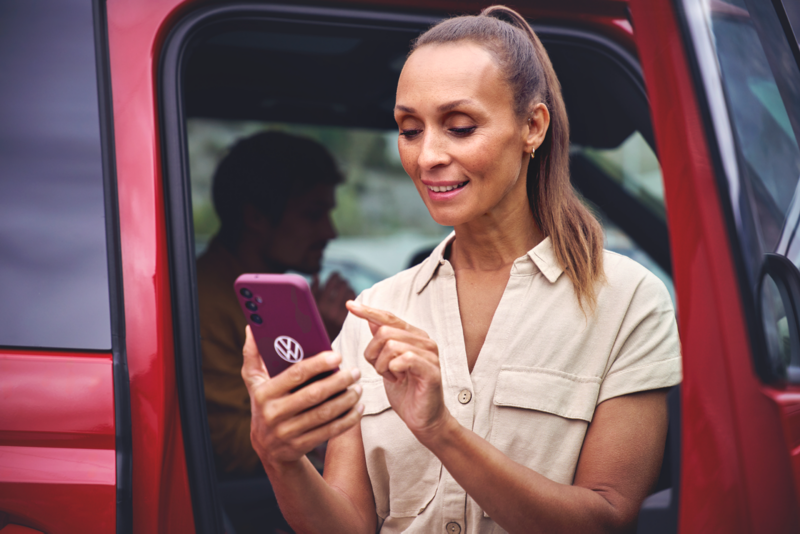 Eine lächelnde Frau steht vor ihrem VW California Campervan und bedient ihr Smartphone.