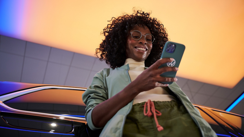 Eine junge Frau in grüner Jacke lehnt an einem Volkswagen Modell und schaut auf ihr Smartphone in der Hand.