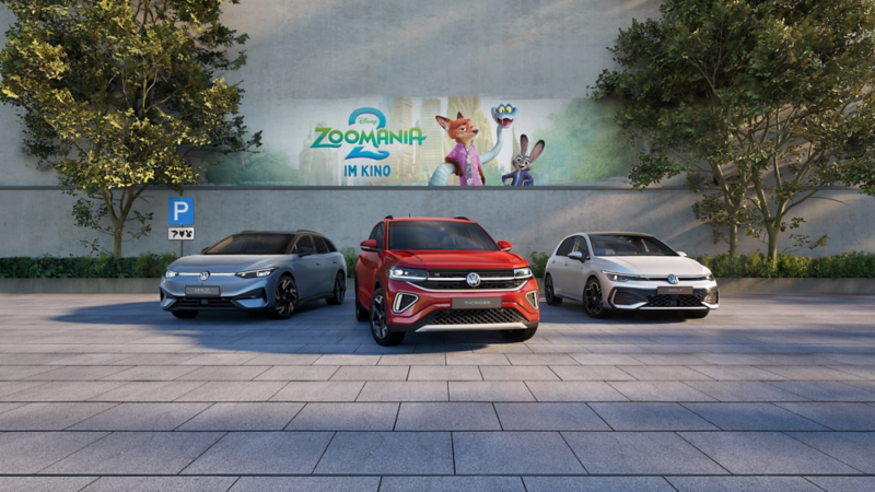 Drei Volkswagen Modelle parken vor einer Betonmauer. Im Hintergrund ist das Motiv des Kinofilms Zoomania 2 zu sehen. 