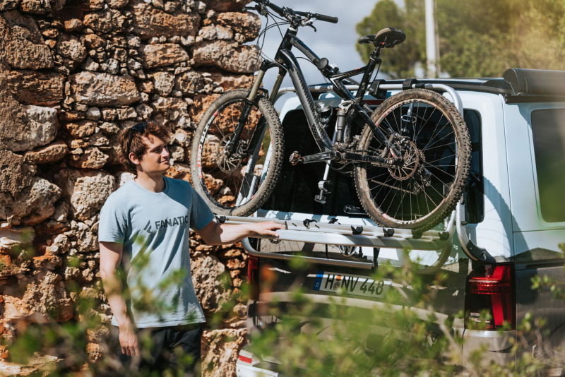 Mężczyzna stoi obok kampera Volkswagen, z rowerem na bagażniku, przymocowanym z tyłu pojazdu.
