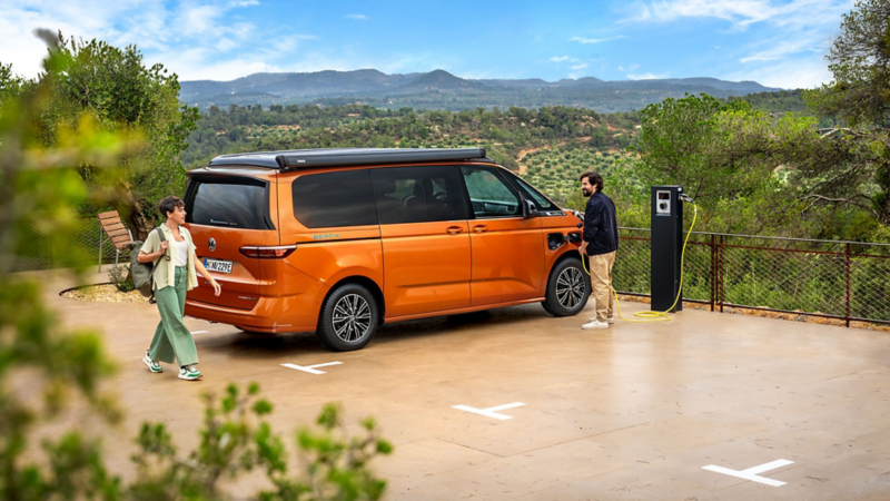 En orange VW California står parkerad vid en laddstation där en person sätter i laddkabeln