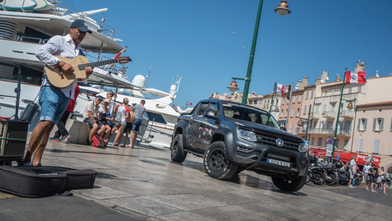 En grå VW Amarok generation 3 står parkerad i en hamn bredvid en yaht och en gatumusiker