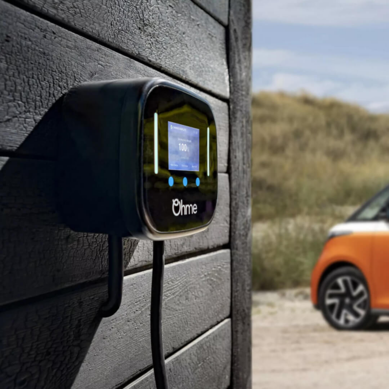 An Ohme Home Pro EV smart charger displaying a 100% charge status is mounted on a dark wooden wall, with an orange electric van blurred in the background.
