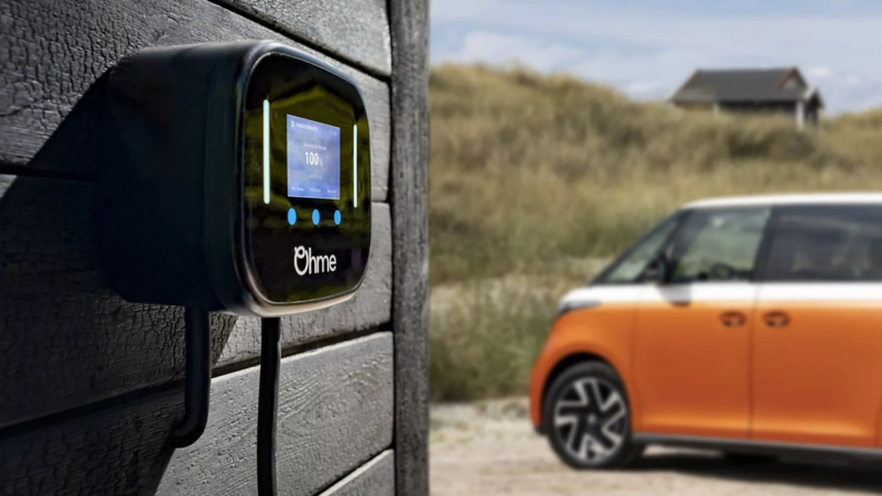 An Ohme Home Pro EV smart charger displaying a 100% charge status is mounted on a dark wooden wall, with an orange electric van blurred in the background.