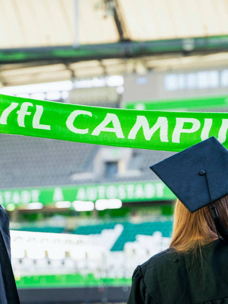 Zwei Absolventen halten im VfL Stadion einen Schal mit Aufschrift VfL Campus hoch.