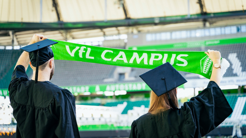 Zwei Absolventen halten im VfL Stadion einen Schal mit Aufschrift VfL Campus hoch.