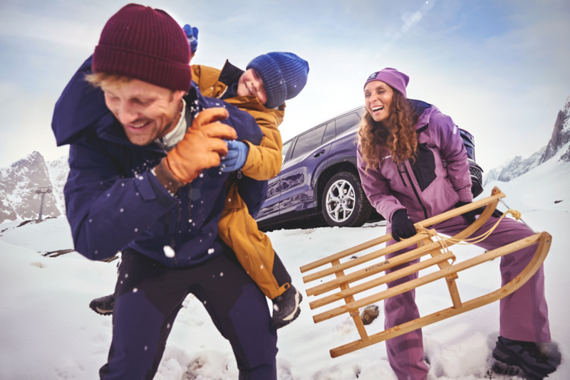 Une famille sourit et s'amuse dans un paysage enneigé. Le père porte un enfant sur son dos, pendant que la mère tire une luge en bois. Une voiture Volkswagen est garée derrière eux.