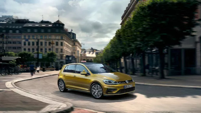 Une voiture Volkswagen Golf dorée roule sur une route sinueuse en ville, bordée d'arbres et de bâtiments.