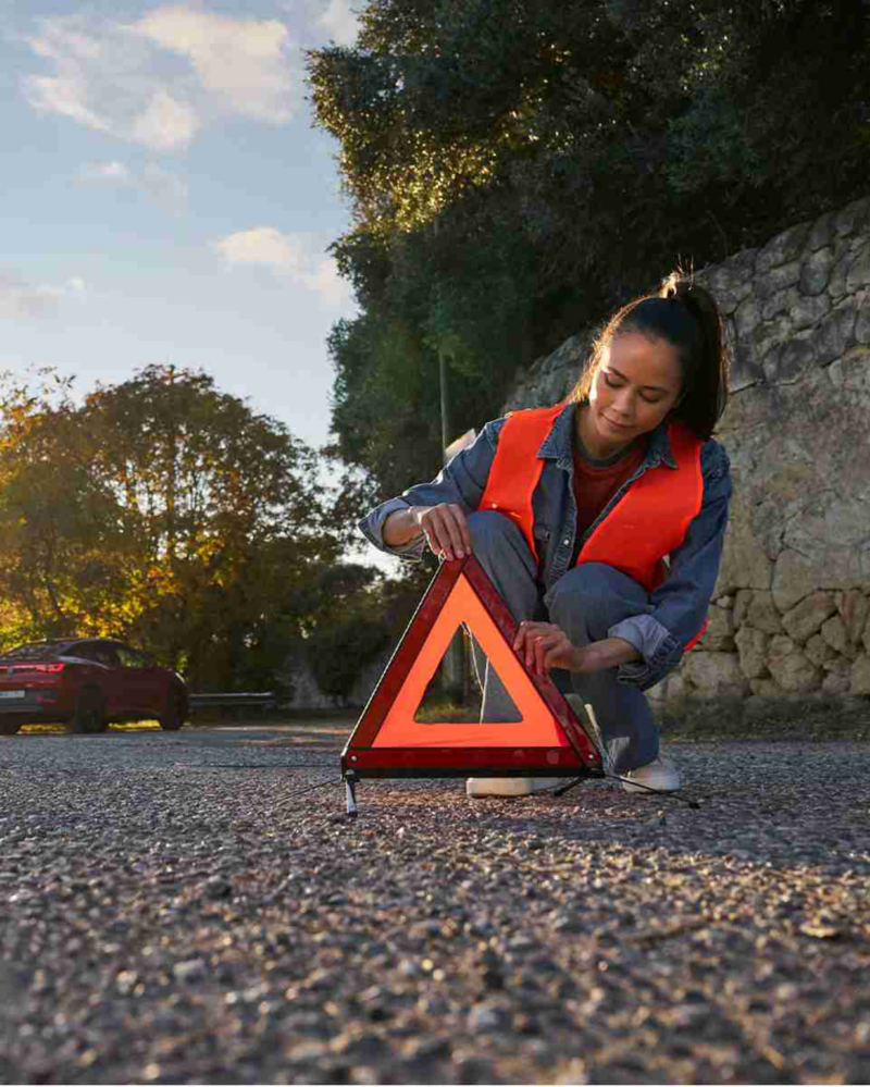 A woman in an orange safety vest crouches to adjust a red warning triangle on the road, with a parked red car and trees in the background.