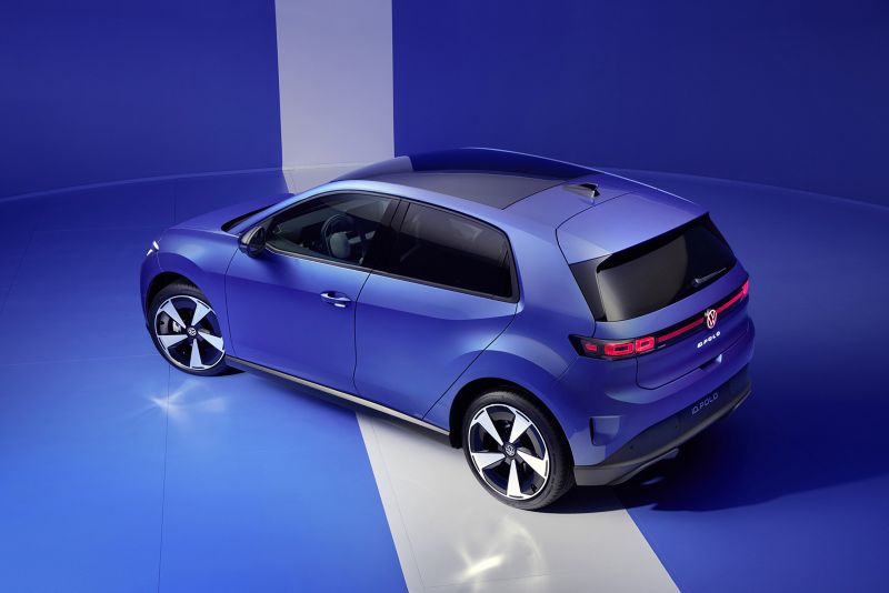 High-angle rear view of a blue Volkswagen ID. Polo electric car parked on a white floor stripe in a blue studio, showcasing its panoramic glass roof and rear design.