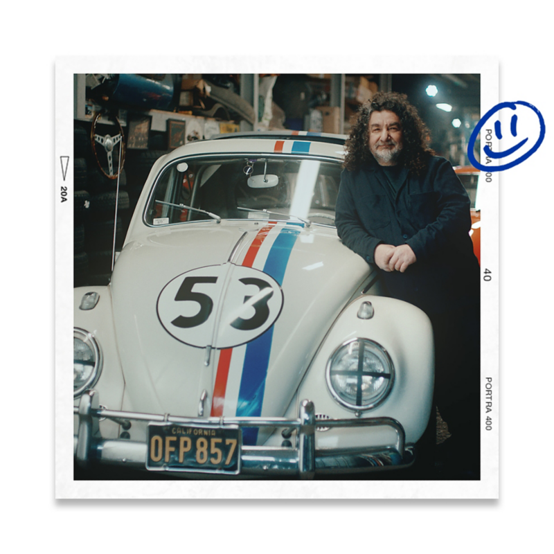 a man with long curly hair leaning on his vintage white VW Beetle