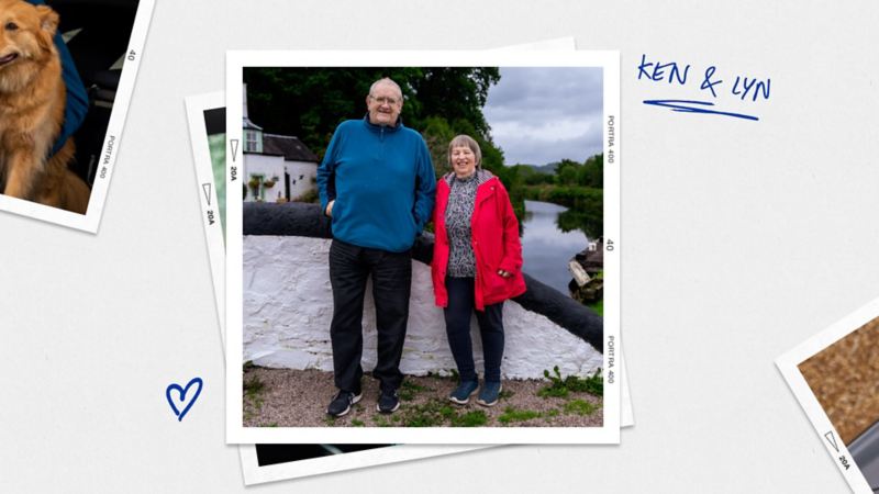 A couple leaning on a wall standing next to a river with a cottage in the background