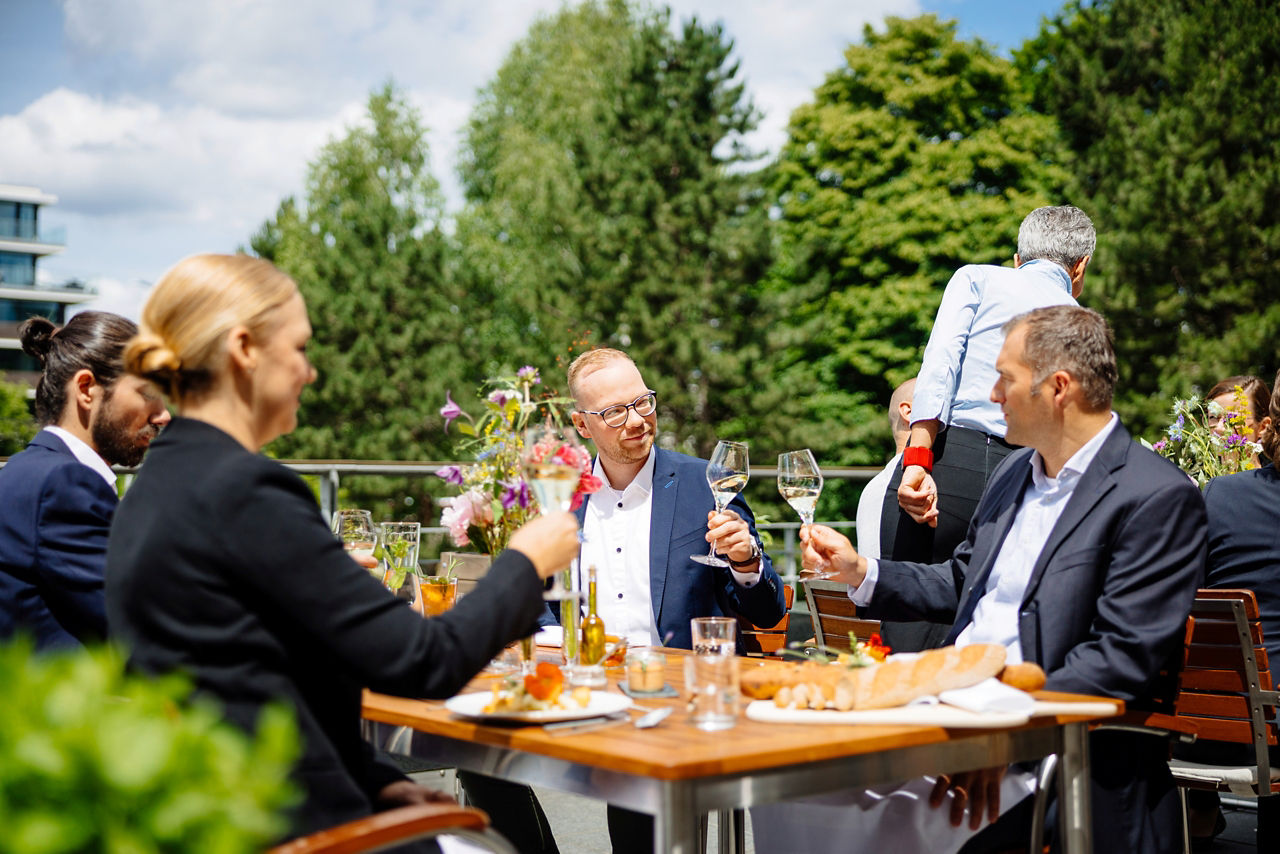 4 Personen sitzen an einem Tisch auf der Terrasse der Manufaktur und stoßen an