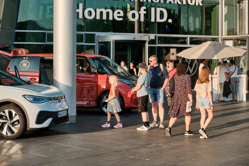Ein gelb-weißer ID. Buzz mit einer geöffneten Seitentür. Zu sehen ist ein Logo der Gläsernen Manufaktur auf der Tür mit der Aufschrift "Happy Sunday"