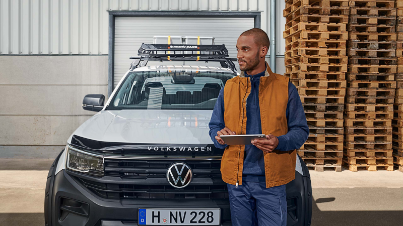 Mann mit Tablet in der Hand steht vor VW Nutzfahrzeug, im Hintergrund Holzpaletten und Rolltor