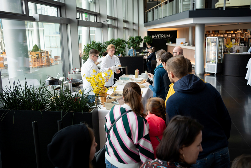 eine Personengruppe steht an einem Tresen. Zwei Herren in weißer Kleidung reichen Teller mit Essen. Im Hintergrund ist der Schriftzug e-Virtum zu lesen