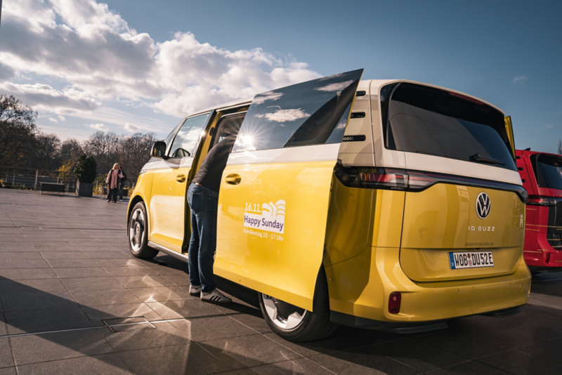 Ein gelb-weißer ID. Buzz mit einer geöffneten Seitentür. Zu sehen ist ein Logo der Gläsernen Manufaktur auf der Tür mit der Aufschrift "Happy Sunday"