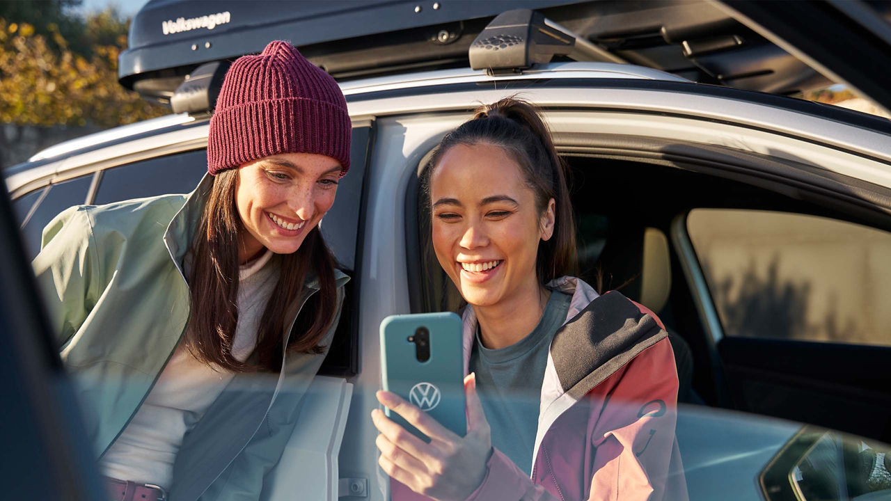 Zwei Frauen schauen auf Smartphone, im Hintergrund ein VW Auto mit Dachbox