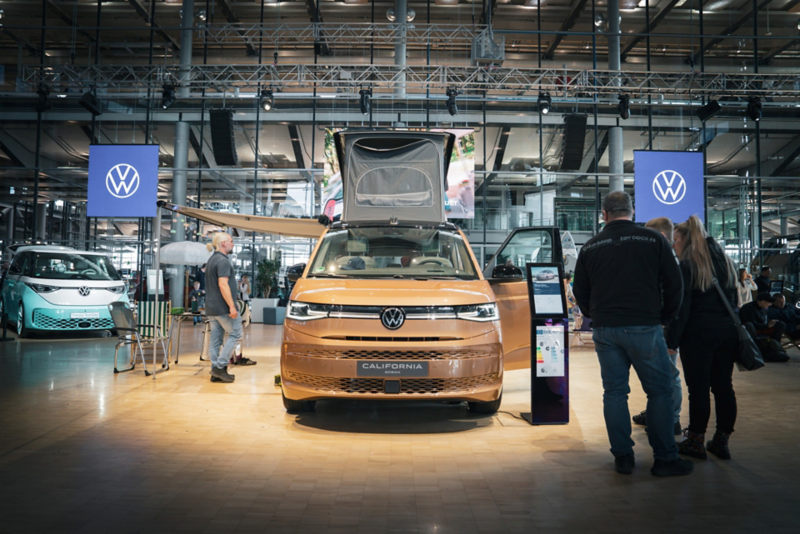 Im Mittelpunkt ein Volkswagen Multivan California mit aufgestellten Dach. Auf zwei Bildschirmen ist ein großes VW Logo zu sehen.