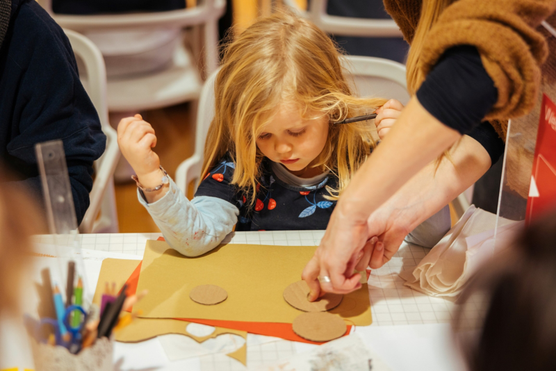 Ein Kind sitzt an einem Platz und hat Bastelutensilien vor sich. Dabei sind zwei weitere Hände zu sehen, die das Kind beim Basteln unterstützen.