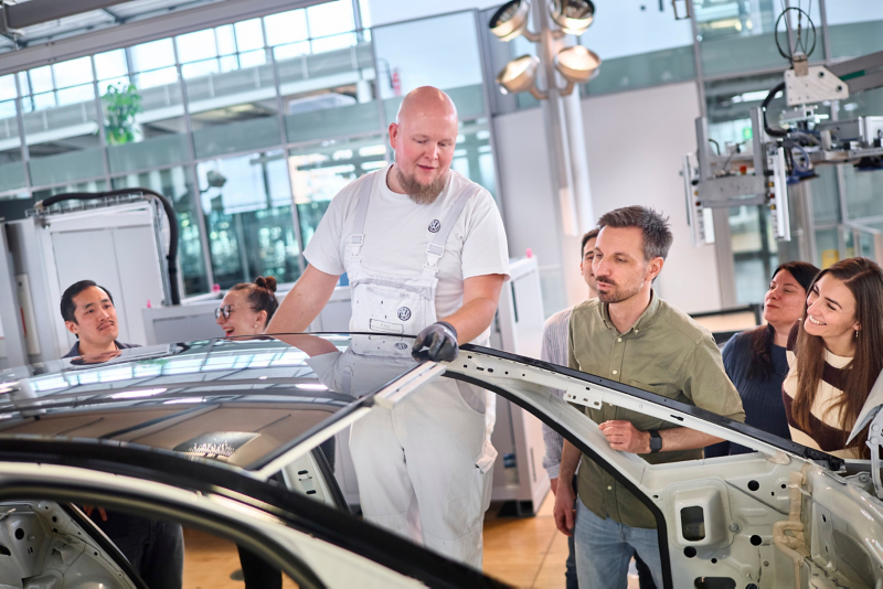 Ein Mitarbeiter seht an einer ID.3 Karosse und prüft den Einbau des Panoramadaches. Eine Besuchergruppe schaut dabei zu.